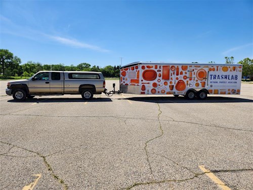 A grey pickup truck towing the Trash Lab - a colorful white and orange trailer - in a parking lot with a clean blue sky in the background.